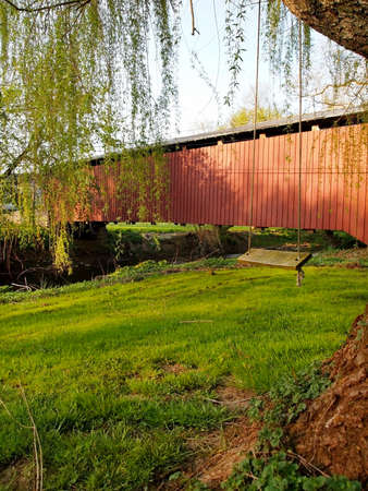 A tree swing hangs from a willow tree near a red covered bridge over a stream in the springtime.