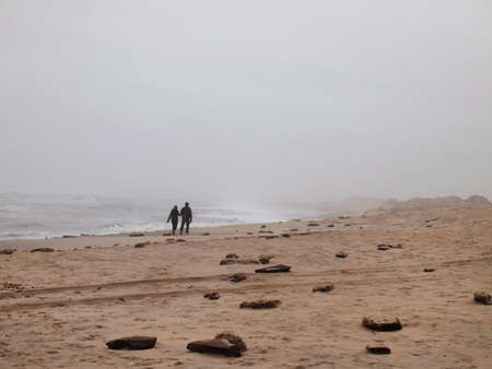 A couple walks hand in hand into the distance on a foggy beach by the water's edge.