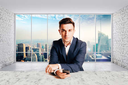 Attractive businessman in trendy suit sitting at desk against panoramic office view backgroundの写真素材