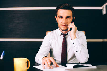 Attractive business man in smart casual wear sitting at his working place in office holding a smart...
