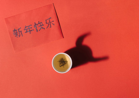 A Chinese tea cup with green tea on red surface. Red lucky envelope with "Happy New Year" greeting in Cantonese characters. Shadow in shape of ox horns 2021 symbol of Lunar New Year.の写真素材