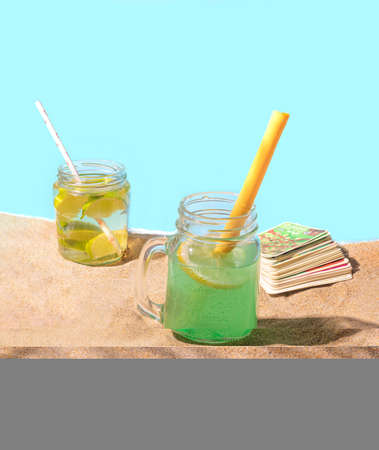 Two jars with cocktail like beverage and paper and reusable silicone straws on the sandy beach, palm leaf shadow, and playing card is the back. Eco friendly concept for environmentally safe vacationの写真素材