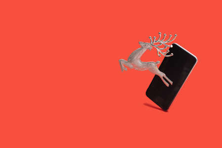 Christmas lovely silver reindeer ornament beside smartphone. Bright red minimal background.