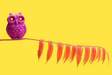 An purple owl glitter toy bird with yellow eyes sitting alone on a colorful leaves tree branch. Bright yellow background.の写真素材