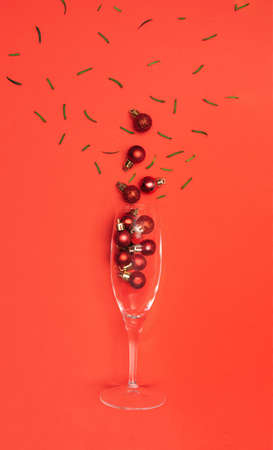 Festive season inspired concept. Champagne glass, green spruce leaves with many shiny red bulbs. Flat lay arrangement with red backgroundの写真素材