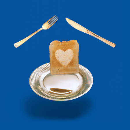 Arrangement made of levitating golden color fork, knife and plate with a slice of toasted bread with a heart shaped in the middle. Modern dark blue background.の写真素材