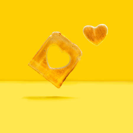 Love is in the air concept. Levitating slice of toasted bread with heart shape inside of it and little heart shaped toast beside it. Bright yellow background.の写真素材