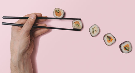 Flat lay sushi vegetarian concept. Man hand holding black chopsticks grabbing one piece, salmon fish, rice, cucumber and nori seaweed between many others different sushi rolls. Pastel pink backgroundの写真素材
