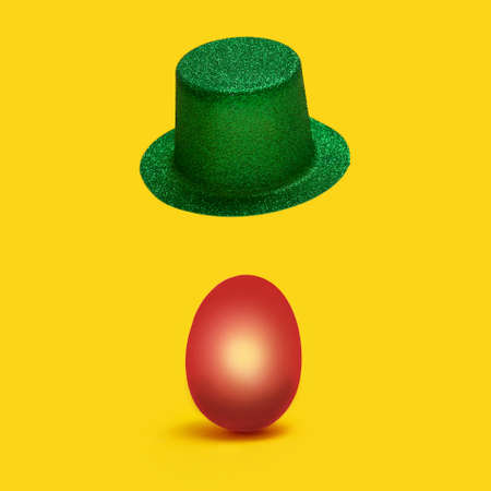 Unique easter minimal concept. Optimistic green hat levitate above red colored egg against yellow backgroundの写真素材