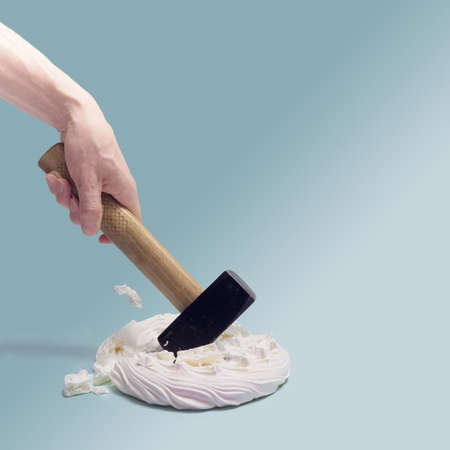 A man hand holding hammer smashed  sweet meringue Pavlova cake. Sky blue background.の写真素材