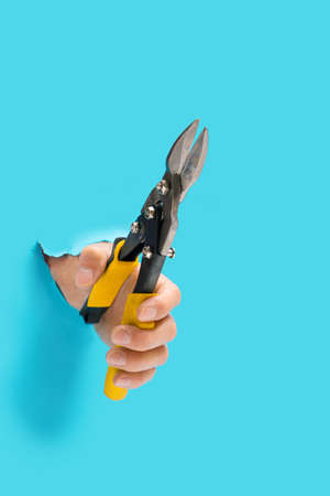 Man hand holding straight cut pliers suitable for cutting plastic or metal materials that breaks through blue wall color background. Aviation snips.の写真素材