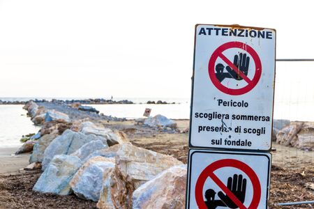 Warning signs reading: 'Warning. Danger submerged reef, rocks on the shoal are present' near a construction site on the shoreline in Follonica, Italyの写真素材