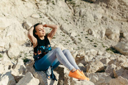Young woman posing near the beautiful mountains dressed colorful clothes sitting on a stoneの写真素材