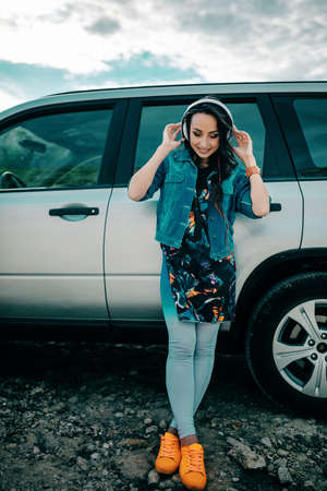 Young woman with headphones posing near car wearing colorful clothes. Sunlight effect