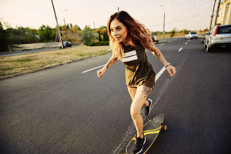 Beautiful young girl with tattoos riding on his longboard on the road in the city in sunny weather. Extreme sportsの写真素材