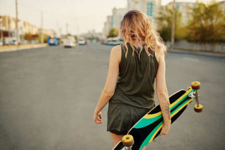 Beautiful young tattooed woman with his longboard on the road in the city in sunny weather