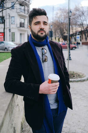 Handsome young man wearing sunglasses on a walk in town. Dreamful guy is relaxing with hot drink