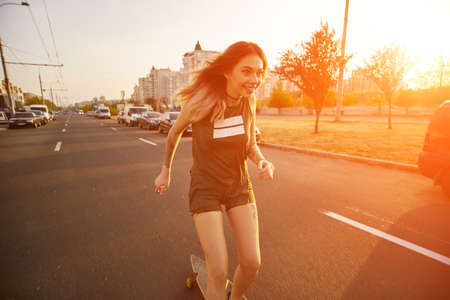 Beautiful young girl with tattoos riding on his longboard on the road in the city in sunny weather....