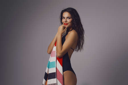 Sexy attractive brunette woman posing in black fashionable swimsuit in studio holding colorful towelの写真素材