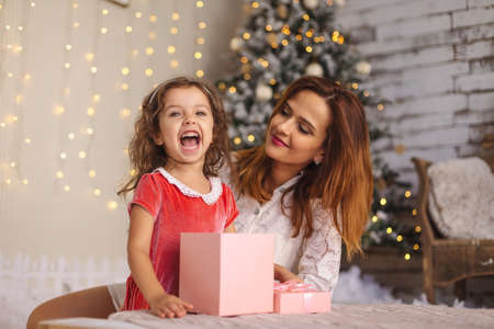 Mother with happy daughter holding Christmas gift box. Cheerful toddler girl looking to camera with open mouth. Little girl open pink gift boxの写真素材