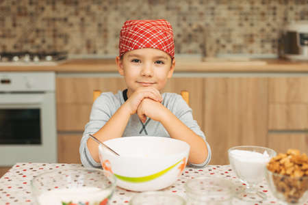 Portrait cute boy shef looking to camera while cooking, sitting in the kitchen at home with ingredients for cake, biscuits on the table.の写真素材