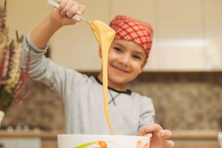 Smiling boy cooking dough for cake having fun, holding a spoon with mixed mas up the kitchen at home, looking to camera, focus on the dough.の写真素材
