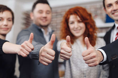 Team holding thumbs up. Close-up of business team holding their thumbs up. Four hands showing thumbs up against the backdrop of the office, two men and two women.の写真素材