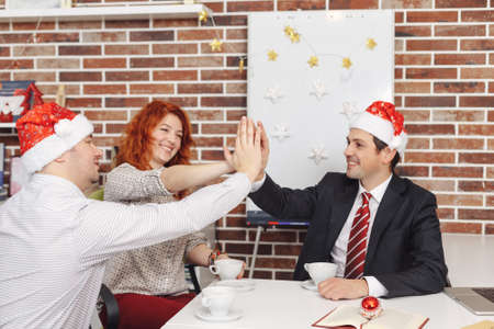 Celebrating Christmas at work. Friendship concept and teamwork. Colleagues drinking tea and give hi five.の写真素材