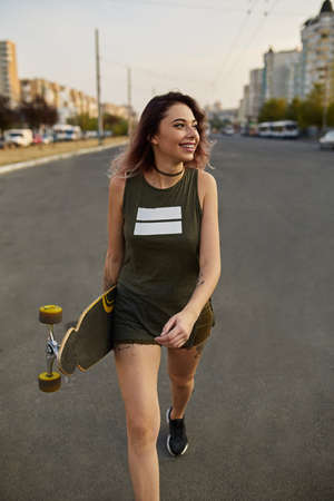 Careless beautiful girl with tattoos posing with a colorful longboard while walking on the road in...