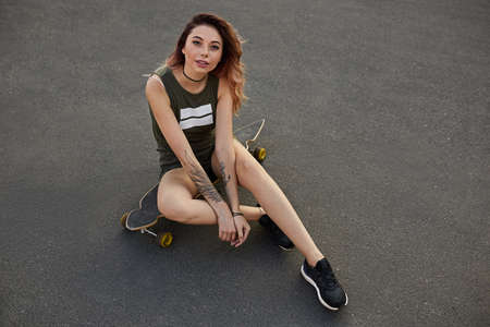 Sexy young girl in short shorts sitting on longboard on the city road in sunny weather. Leisure. ...