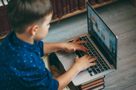 Angle view, boy sitting on the floor with his feet crossed, typing on a laptop against bookshelf in library. Education, Knowledge, Bookstore, Lecture. laptop on a stack of books.の写真素材