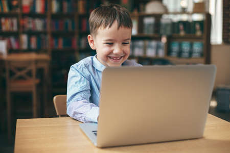 Smiling preschool boy is focusing on his laptop, E-Learning concept. Digital composite of  boy at table looking at a computer against books in a library.の写真素材