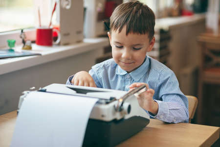Smiling preschool journalist boy sitting at table and typing typewriter with  a pencil on the top of year. Businessman using typewriter at desk in office. Business and new technology.の写真素材