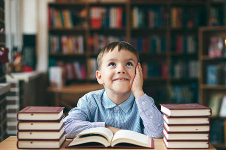 Single dreaming boy at the desk in the library enjoy books. Front image with boy keeping left hand on the cheek . European boy explore books. Pupil loves lecture, education, preparing for school.の写真素材