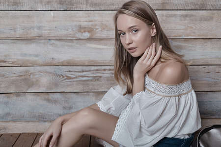 Beautiful girl wearing natural makeup, linen dress and boots. Concept of purity, youth, beautiful skin sitting on pastel wood background, with a silver backpack, touching face while looking to camera.の写真素材