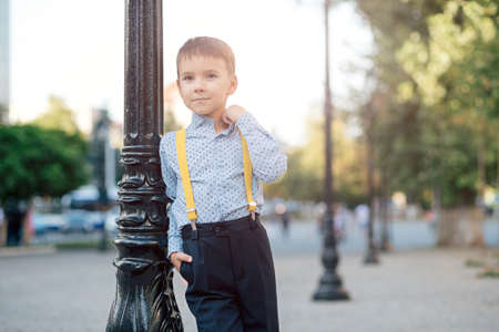 Fashion full length boy portrait in dark blue pants, baby blue shirt and yellow straps, standing on pedestrian street with arranging shirt collar looking to camera, urban background.の写真素材
