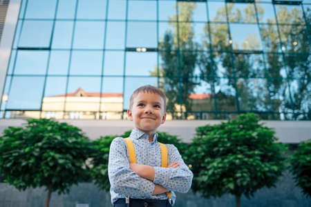Boy portrait, down angle, future businessman, wearing yellow suspenders, blue shirt against office building with round trees, with crossed arms looking to camera.の写真素材