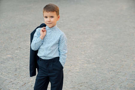 Portrait elegant little man in fashionable navy blue shirt holding dark blue jacket on his back smiling and looking to camera on urban gray pavage. Young boy posing. Boy ready for school.の写真素材