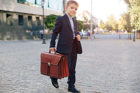 Concept future businessman. Business meeting. Low angle of future businessman in formal wear walking around the street carrying a braun briefcase and holding jacket with arm, urban background.の写真素材