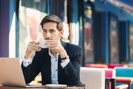 Young entrepreneur working on laptop, at the table on colorful cafe terrace and enjoying coffee holding the cup in his hands. Freelancer, in casual jacket, unbuttoned shirt thinking on future plans.の写真素材