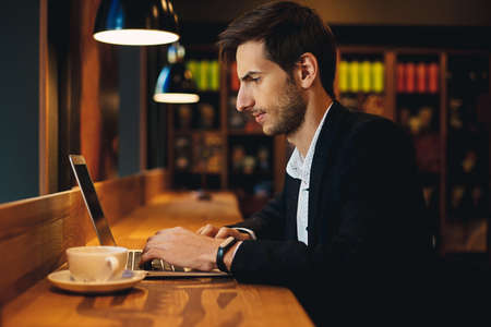 Young entrepreneur working on laptop sitting at wooden table in a modern cafe. Freelancer, in casual black jacket and unbuttoned shirt typing while looking to laptop screen, with a cup of coffee.の写真素材