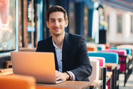 Smiling modern entrepreneur working on laptop sitting at the table in a colorful cafe. Ambitious freelancer, dressed in casual black jacket and unbuttoned shirt typing while looking to camera.の写真素材