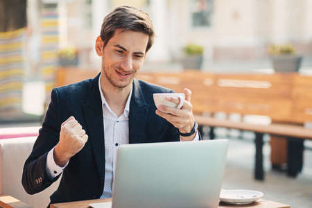 Emotions concept. Guy with winner look sitting at table holding cup of coffee in hand looking to laptop. Young man is happy to have the contract signed, to get the email with job acceptance.の写真素材