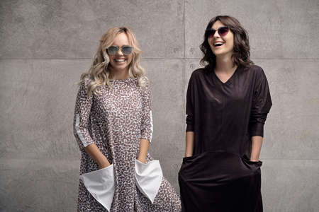Portrait two cheerful fashionable young women wearing sunglasses, dresses and sneakers posing on...