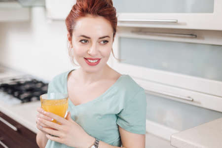 Portrait smiling good looking woman drinking a glass of orange juice while standing in the kitchen.の写真素材
