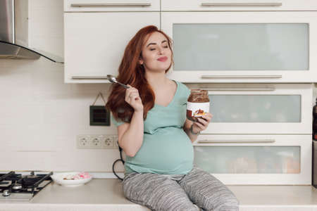 Beautiful pregnant woman enjoying eating chocolate paste. Happy face expresion, closing eyes of pleasure, sitting on the kitchen furniture. Portrait.の写真素材