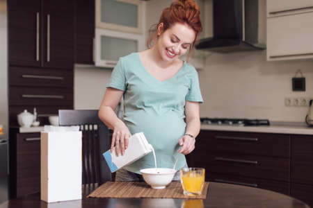 Smiling pregnant woman preparing breakfast, cereals with milk and natural juice, in the kitchen.の写真素材