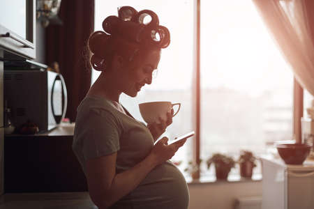 Pregnant woman got good news. Female profile picture while drinking a cup of tea while using smartphone, near the window with city reflection in it. Female silhouette profile picture.の写真素材