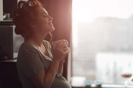 Pregnant woman emotions concept. Living feelings while drinking a cup of tea near the window with...