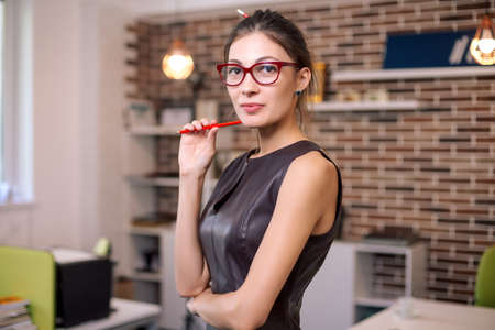Portrait of successful smiling business woman, worker, designer, lady, female wearing glasses and leather dress looking to camera while touching her cheek with a red pencil on office background.の写真素材